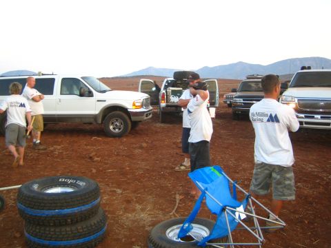 1990 Ford Bronco - People in McMillin Racing shirts stand with stacked Baja spare tires and a camp chair beside pickups and a white truck in a dusty pit.