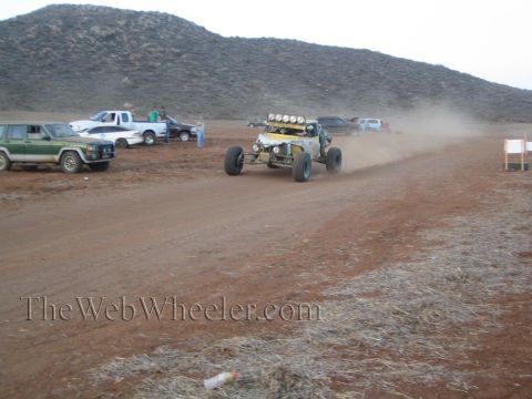 1990 Ford Bronco - yellow off-road race buggy with a roof light bar races on a red-dirt desert course past parked pickups and a green Jeep.