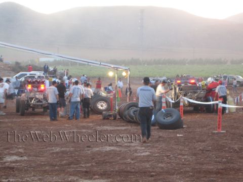 1990 Ford Bronco - busy desert race pit area with crews working on off-road cars, stacked spare tires, cones and pennant tape at dusk.