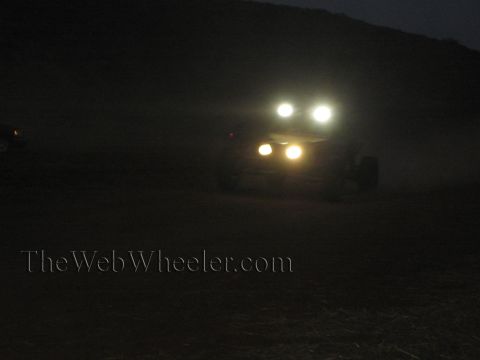 1990 Ford Bronco - off-road race vehicle approaches at night, its headlights and roof light bar glowing through the dark desert dust.