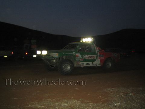 1990 Ford Bronco - green and red off-road race truck with Castrol livery and lit light bar sits in a dark desert with headlights on.