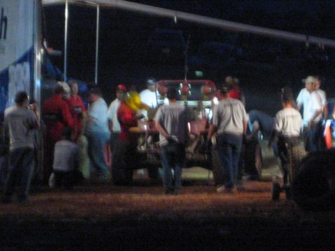 1990 Ford Bronco - crowd of people gathers around a red off-road race car parked beside a hauler trailer in a dark desert pit at night.