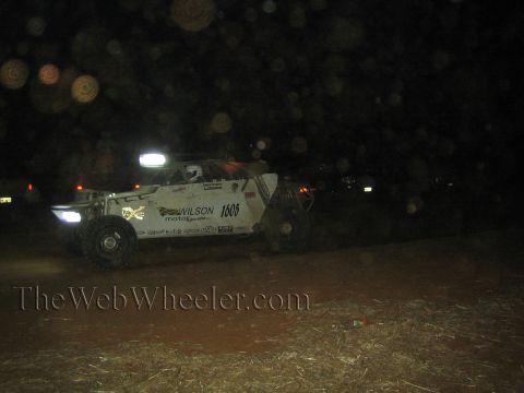 1990 Ford Bronco - White off-road race buggy number 1605 with Wilson Motorsports livery and lit light bar runs across a dark desert course at night.