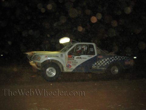 1990 Ford Bronco - white and blue number 85 off-road race truck with checkered-flag graphics and Yokohama decals runs at night with light bar on.