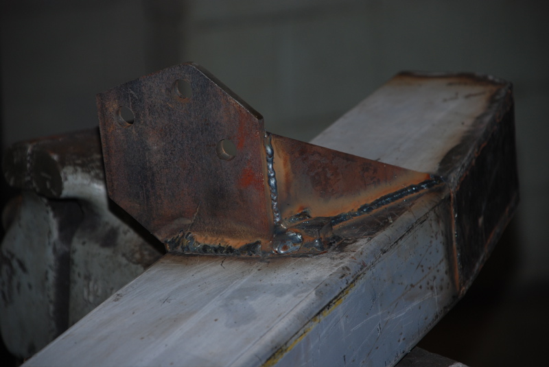 Welded metal bracket attached to a gray beam, showing rust and rough welds