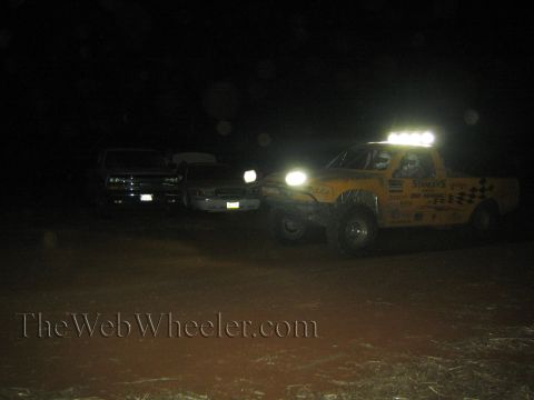 1990 Ford Bronco - yellow off-road race truck with its roof light bar lit and Sammys decals sits beside two parked support trucks at night.