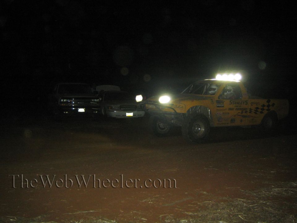 Yellow off-road race truck with light bar beside two parked trucks at night