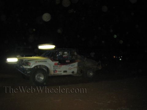 1990 Ford Bronco - white off-road race truck with Mango Racing and Yokohama decals and yellow-tinted light bars runs at night on dirt.