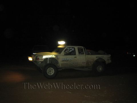 1990 Ford Bronco - muddy white off-road race truck with Looney and BFGoodrich decals runs at night with its roof light bar illuminated.