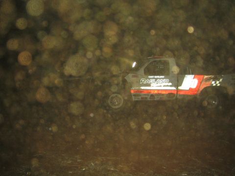 1990 Ford Bronco - white off-road race truck with Ragland sponsor and red graphics and checkered flag races at night amid heavy dust.