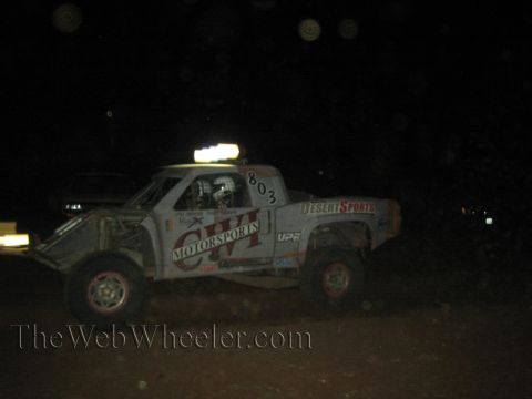 1990 Ford Bronco - white CVI Motorsports number 803 off-road race truck with Desert Sports decals runs at night with light bar lit.