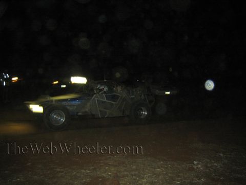 1990 Ford Bronco - dark open-frame off-road race buggy sits on a dirt course at night with spectators and glowing lights around it.