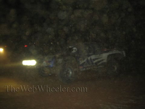 1990 Ford Bronco - off-road race truck speeds past at night with its light bar glowing, partly obscured by dust and lens flare.