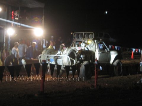 1990 Ford Bronco - white off-road race truck sits in a floodlit pit area at night surrounded by crew and spectators behind safety bunting.