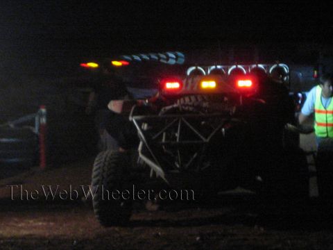 1990 Ford Bronco - rear of an off-road race buggy at night shows glowing red lights, a tube roll cage and a crew member in a safety vest.