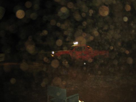 1990 Ford Bronco - red off-road race truck speeds across a dark course at night, its roof-mounted light bar glowing through camera lens flare.