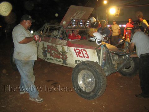 1990 Ford Bronco - man in a ball cap stands beside the open number 1213 off-road buggy as a crew works on it in a dirt pit lit by overhead lights.