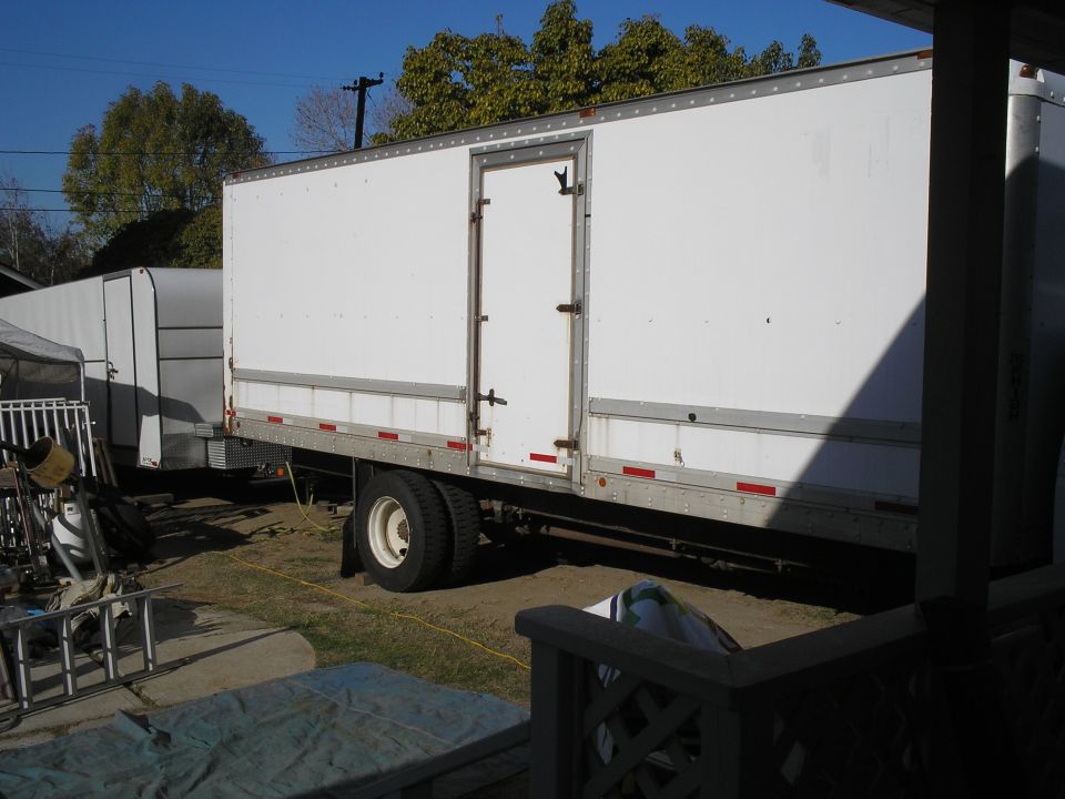 White box truck side view with side door and reflective strips, parked in a dirt yard near a white trailer