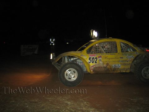 1990 Ford Bronco - yellow VW Beetle-bodied off-road race car marked 502 races across a dark dirt course at night, covered in sponsor decals.