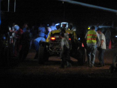 1990 Ford Bronco - rear of a yellow off-road race car glows with brake lights as crew and spectators gather around it in a dark dirt pit area.