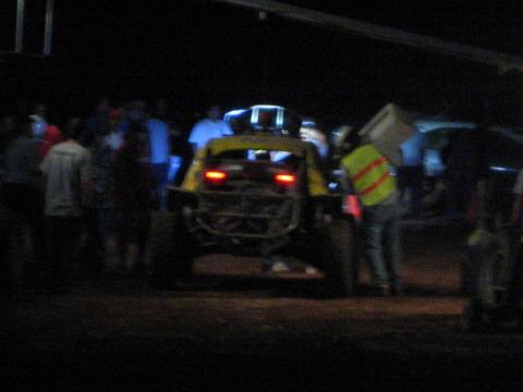 1990 Ford Bronco - yellow off-road race car shows its glowing tail lights as a crowd of people surrounds it on a dark dirt course at night.