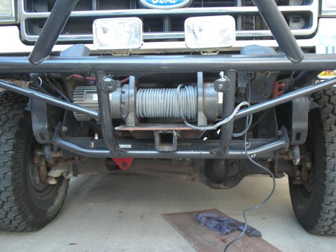 1990 Ford Bronco - Head-on view under the front of a Ford truck showing a fabricated black tube bumper with a winch mounted behind it, its drum wrapped in ste…