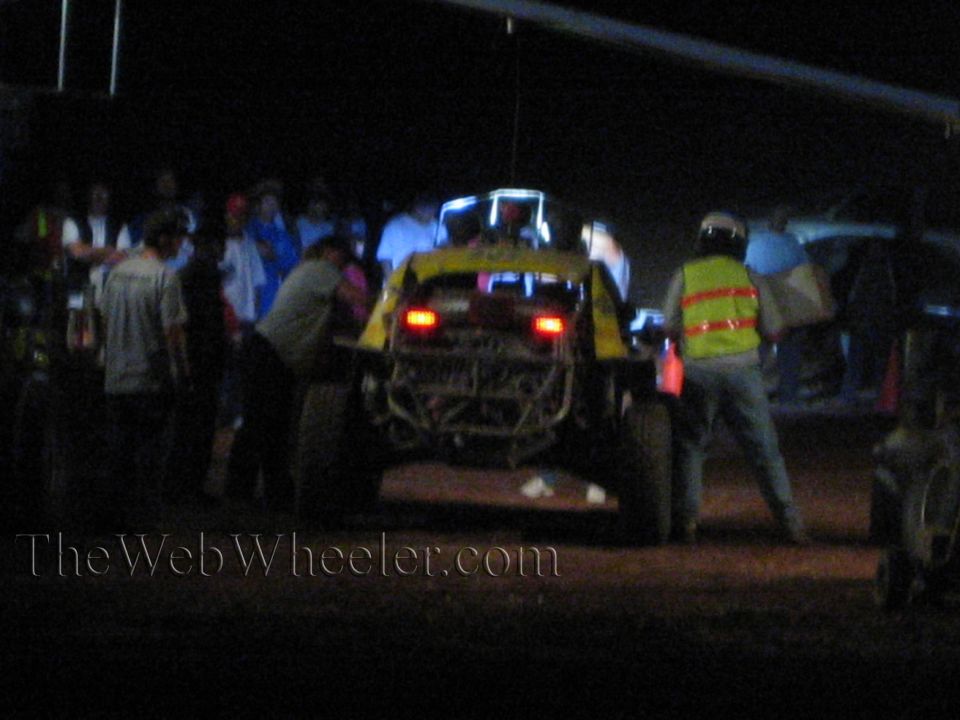 Yellow off-road race car rear with brake lights and pit crew at night