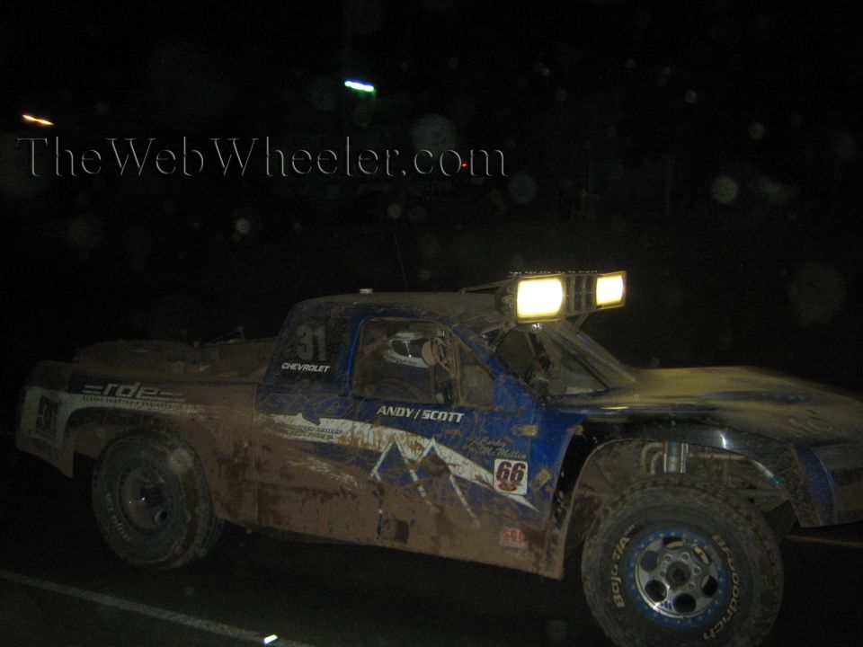 Blue and white muddy off-road race truck with lit roof lights at night