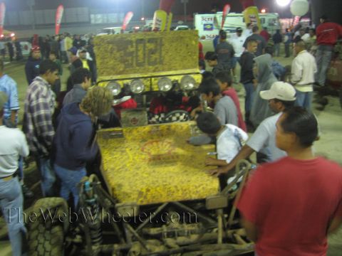 Mud-caked yellow off-road buggy with roof light bar surrounded by a crowd at night