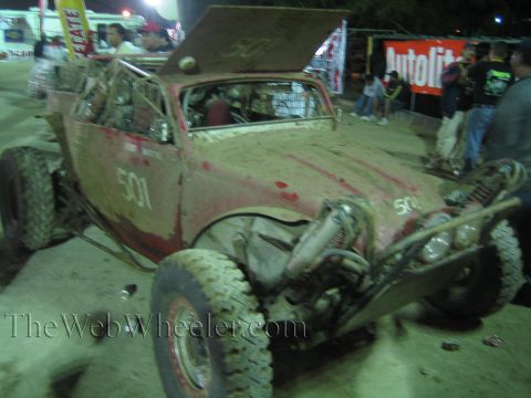 Red dirt-covered off-road car number 501 with exposed engine at a night pit