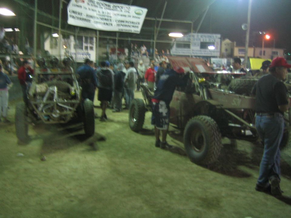 Crowd of people around tube-frame off-road buggies on arena dirt at night