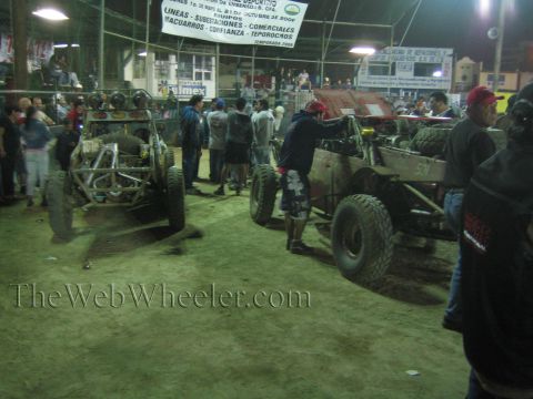 Two tube-frame off-road buggies and a crowd inside a covered arena at night