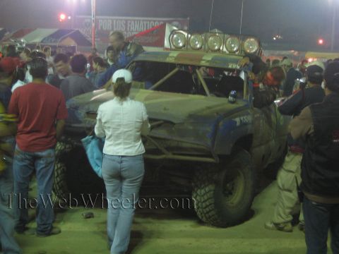 Off-road race truck number 296 with roof lights and a crowd under Por Los Fanaticos banner