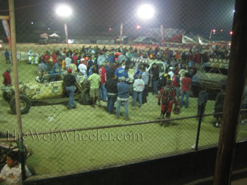 Large nighttime crowd packed among off-road vehicles seen through a chain-link fence