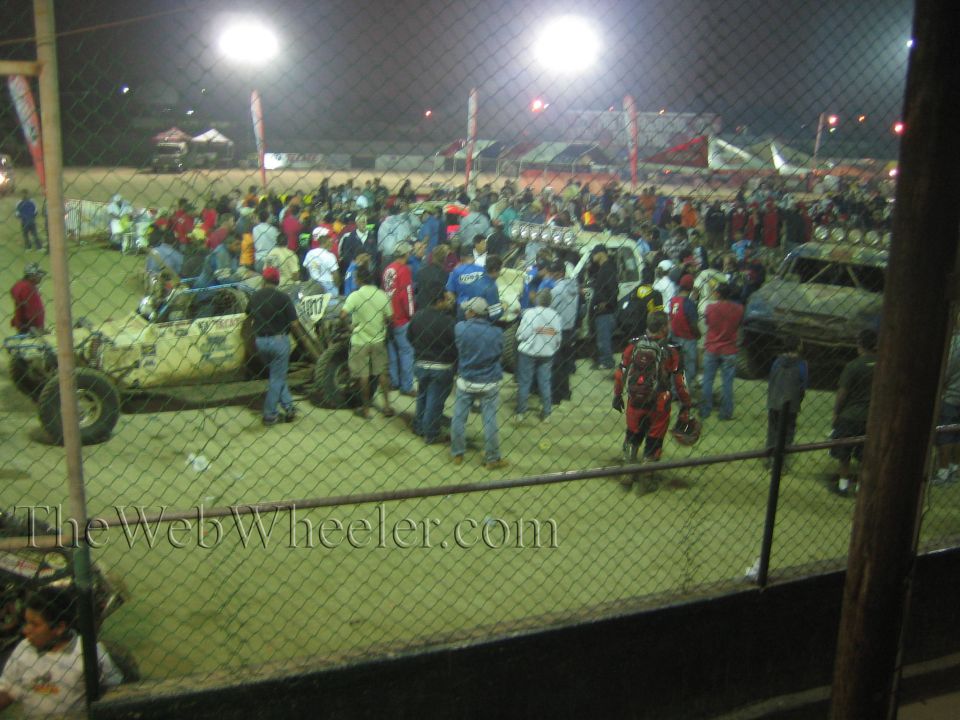 Large nighttime crowd packed among off-road vehicles seen through a chain-link fence