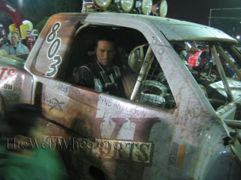 Driver in helmet inside muddy off-road truck number 803 with name decals at night