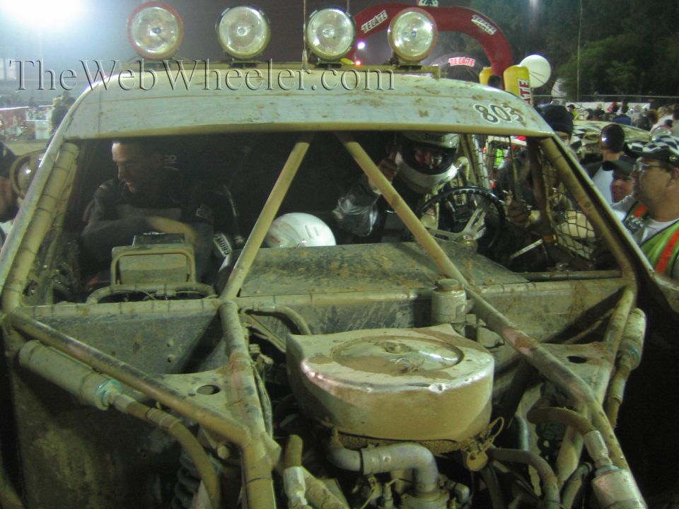 Driver in helmet inside mud-caked off-road truck number 803 with roof light bar
