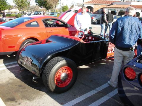 1998 Ford Crown Victoria - Rear three-quarter of a black hot rod roadster with red wire wheels parked beside an orange Mustang at a car meet.