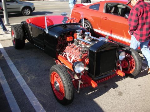1998 Ford Crown Victoria - Black hot rod roadster with a chromed dual-carb V8 and red wire wheels parked beside an orange Mustang at a meet.