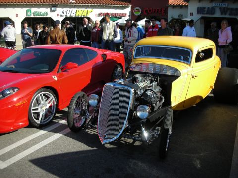 1998 Ford Crown Victoria - yellow hot rod coupe with an exposed chrome engine parked beside a red Ferrari F430 outside storefronts.