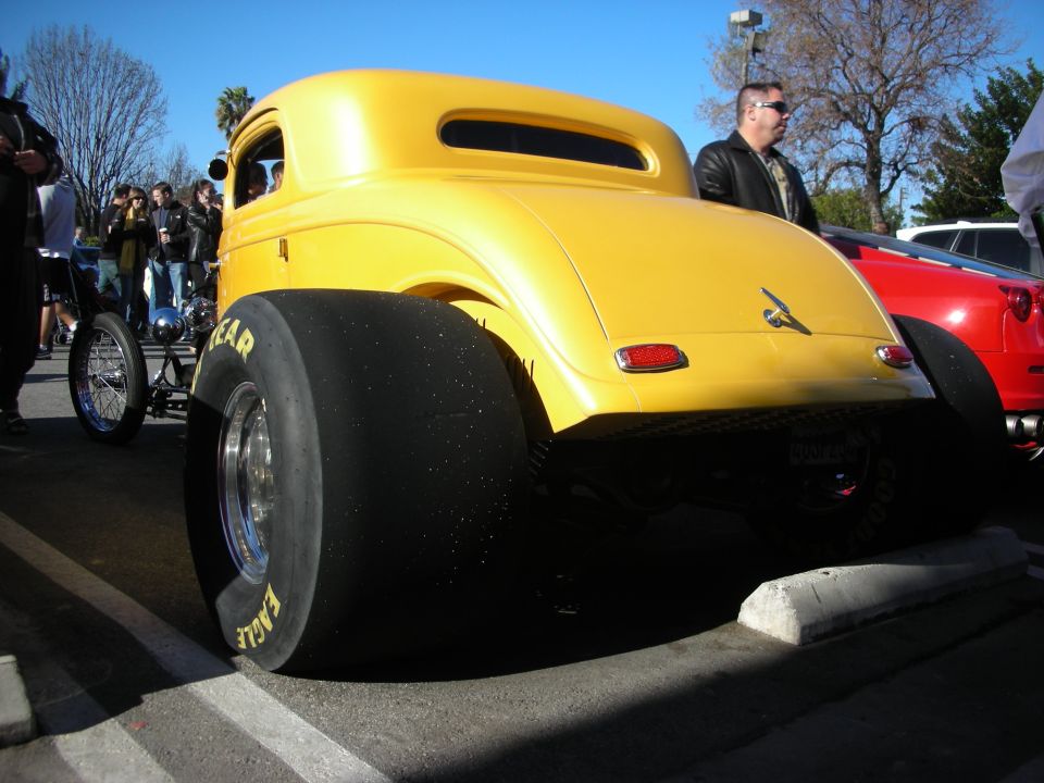 Rear of a yellow hot rod coupe with wide slick drag tires at a car show