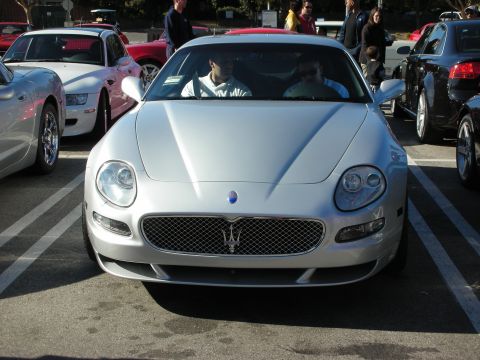 1998 Ford Crown Victoria - silver Maserati coupe seen head-on in a parking lot, its mesh grille and trident badge prominent, surrounded by other cars and onlookers.