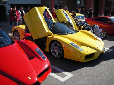 1998 Ford Crown Victoria - yellow Ferrari Enzo with its scissor door up beside a red Enzo, with a vintage car and crowd in the background.