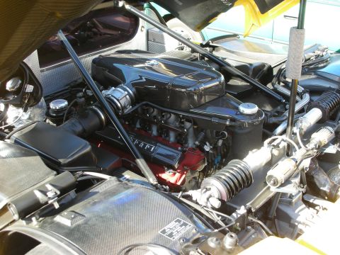 1998 Ford Crown Victoria - open engine bay of a Ferrari revealing a V12 with carbon-fiber covers, red detailing, and exposed suspension links.