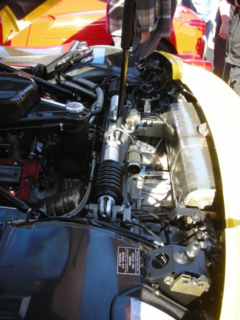 1998 Ford Crown Victoria - open engine compartment of a yellow sports car showing a red cam cover, exhaust manifolds, and a cautionary placard.