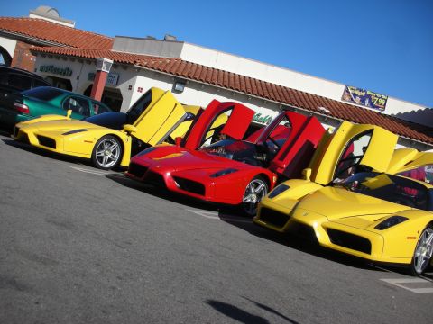 1998 Ford Crown Victoria - row of Ferrari Enzo supercars, two yellow and one red, sit with their dihedral doors raised in a sunny shopping-center lot.