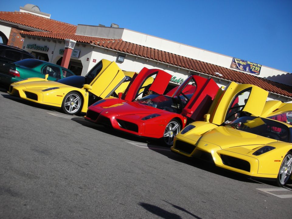 Two yellow and one red Ferrari Enzo supercars parked with doors open at a strip mall