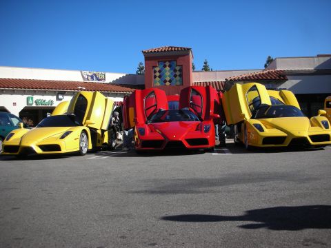 1998 Ford Crown Victoria - Yellow and red Ferrari Enzo supercars line up with doors open before a tiled-roof shopping plaza under a clear blue sky.