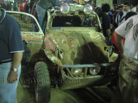 Muddy off-road buggy with roof lights and stickers surrounded by a crowd at night