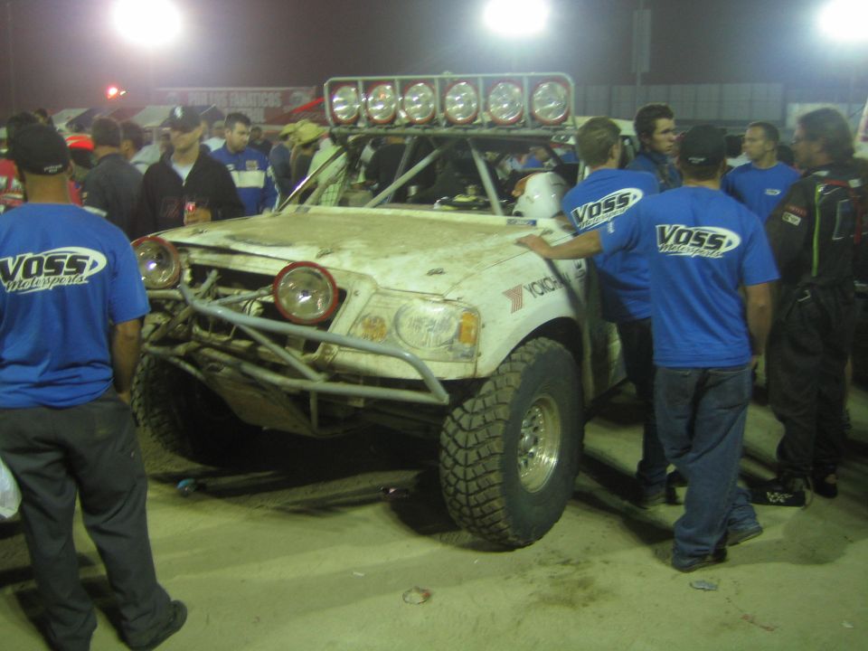 White off-road truck with roof lights surrounded by crew in blue Voss Racing shirts